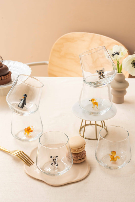 A set of clear drinking glasses with handcrafted animal figures inside, displayed on a table with dessert and tea sets in the background.